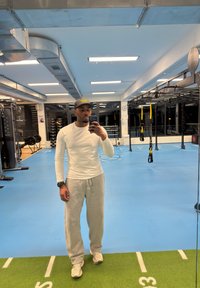 Homme en chemise blanche à manches longues, pantalon de survêtement gris et casquette prenant un selfie dans une salle de sport avec un sol bleu et du matériel d'entraînement.