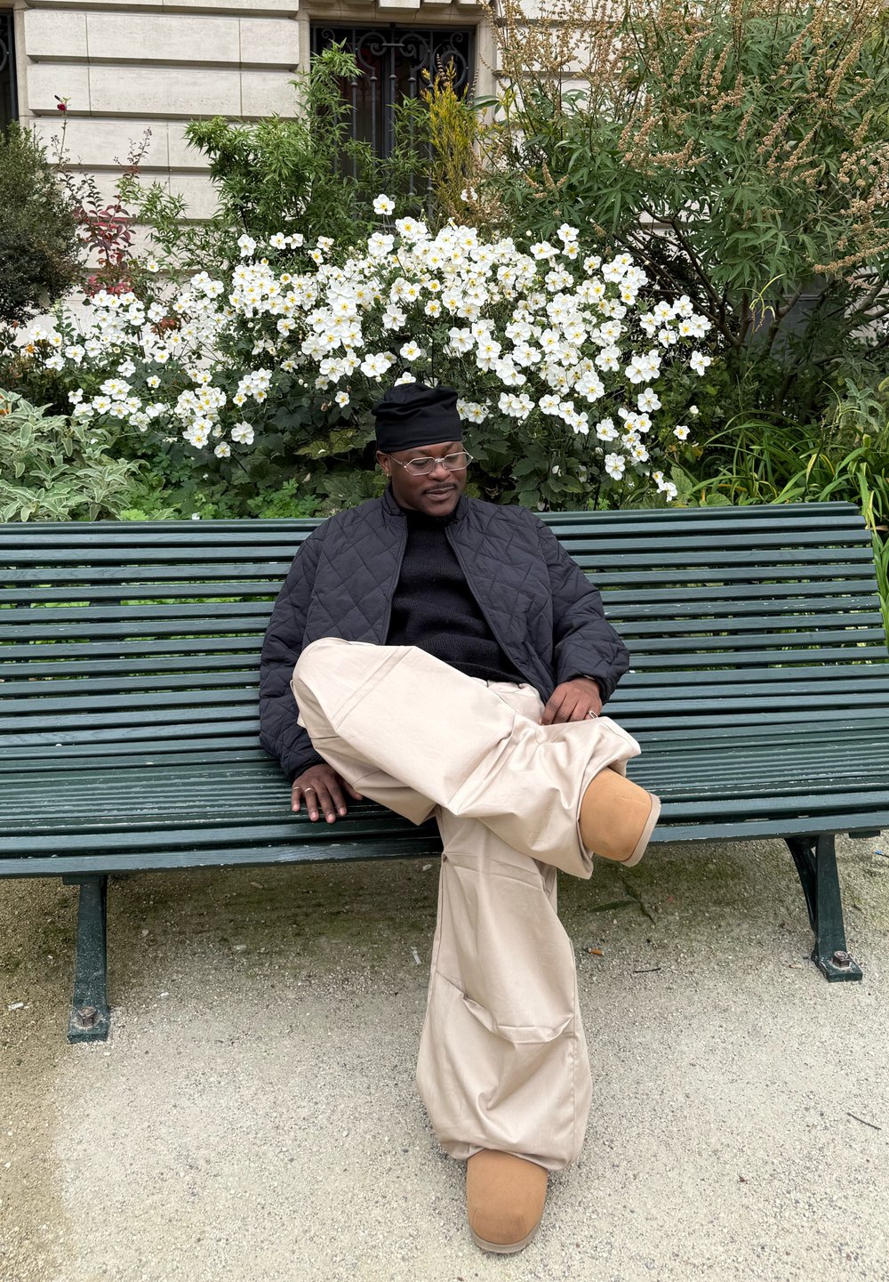 Osoba sediaca na zelenej lavičke, oblečená v čiernej prešívanej bunde, čiernom svetri a béžových nohaviciach s širokými nohavicami spolu s hnedou obuvou. Kvetinové pozadie.