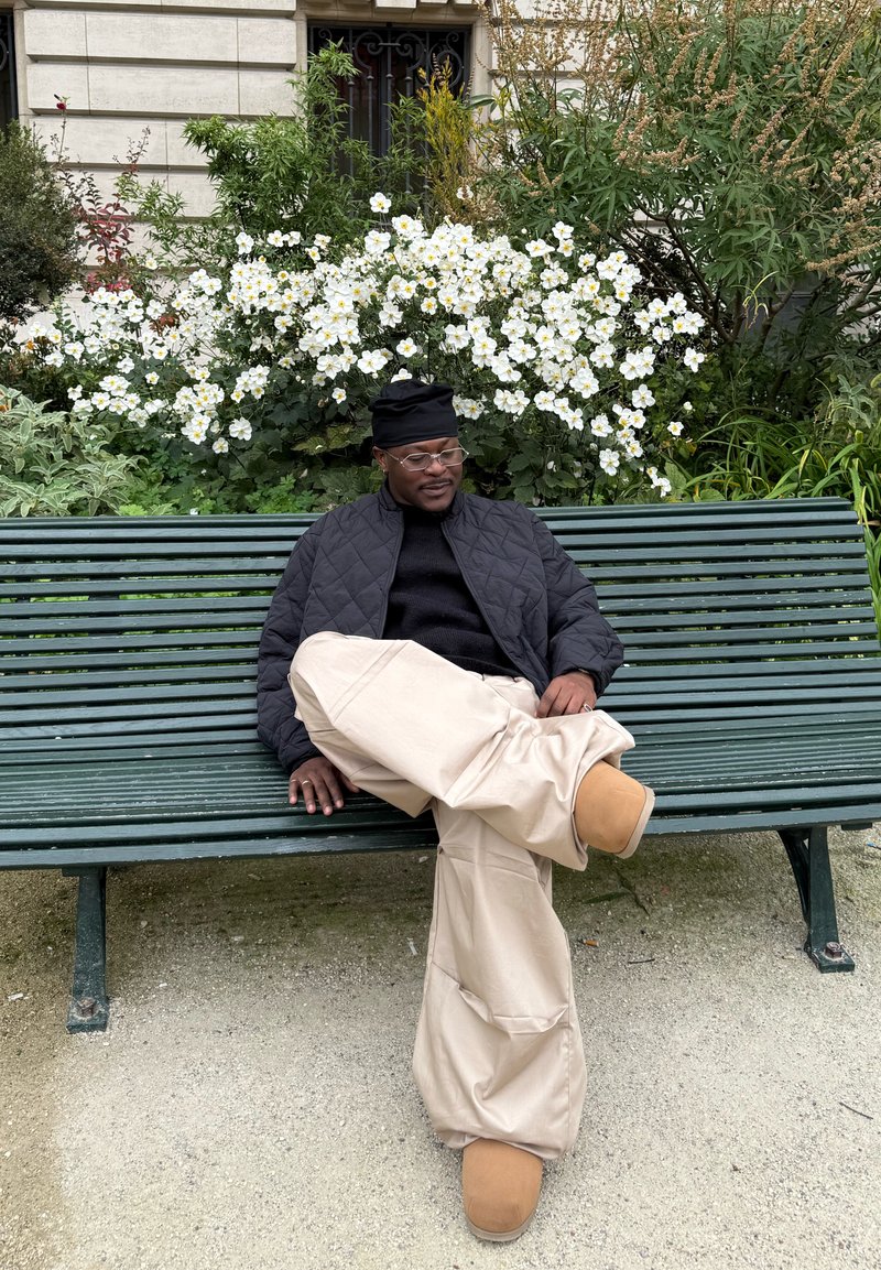 Personne assise sur un banc vert, portant une veste matelassée noire, un pull noir et un pantalon beige à jambes larges avec des chaussures marron. Fond floral.