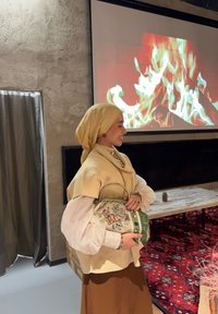 Femme portant un foulard beige et un chemisier, tenant un sac brodé, souriant à côté d'une table et d'un écran affichant une cheminée en feu.