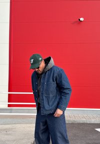 Veste en denim et pantalon assorti en bleu foncé, superposés avec un sweat à capuche gris ; portant une casquette verte avec des détails blancs. Fond rouge.