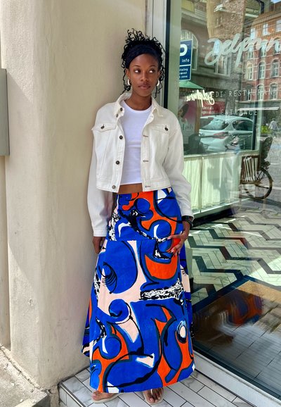 Chaqueta de denim blanca sobre un top corto blanco, combinada con una falda larga con atrevidos patrones abstractos en azul, naranja y negro. Sandalias planas.
