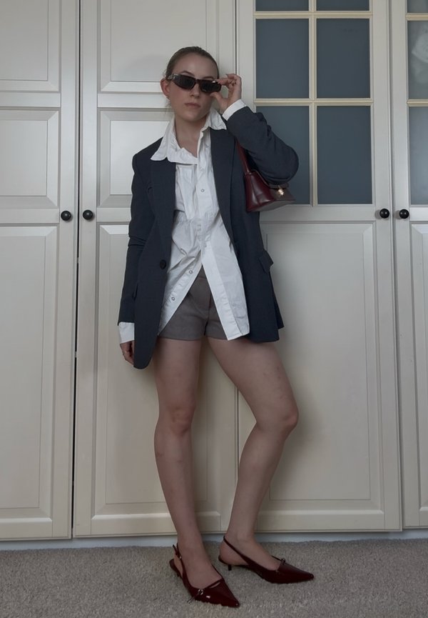 Mujer de pie en el interior con gafas de sol, blazer oscuro de gran tamaño, camisa blanca, pantalones cortos grises, bolso rojo oscuro y tacones puntiagudos a juego.