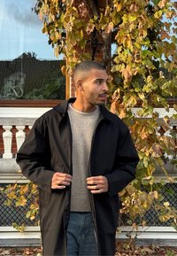 Sort, let yderjakke med krave og sænkede skuldre; båret over en grå striktrøje. Baggrunden viser efterårsblade.