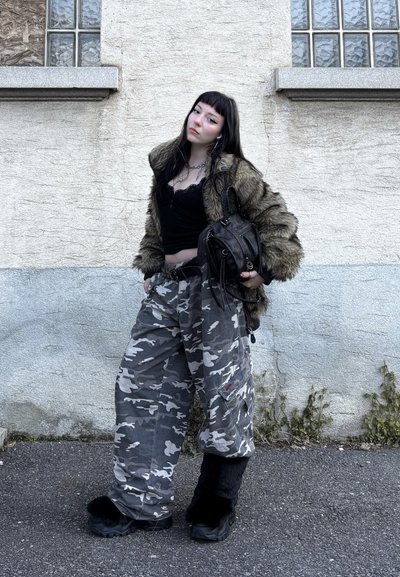 Chaqueta de piel sintética verde oliva, top negro, pantalones cargo gris con estampado de camuflaje y botas negras peludas. Los accesorios incluyen joyas en capas y un bolso negro.