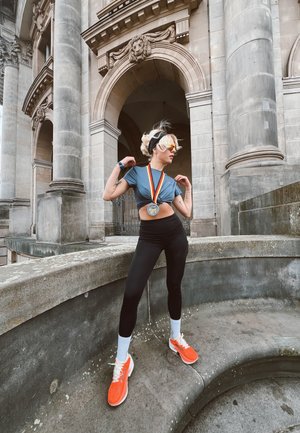 Joven atleta luciendo una medalla, zapatillas de correr naranjas, leggins negros y un top corto azul, posando junto a arcos y columnas de piedra históricas.
