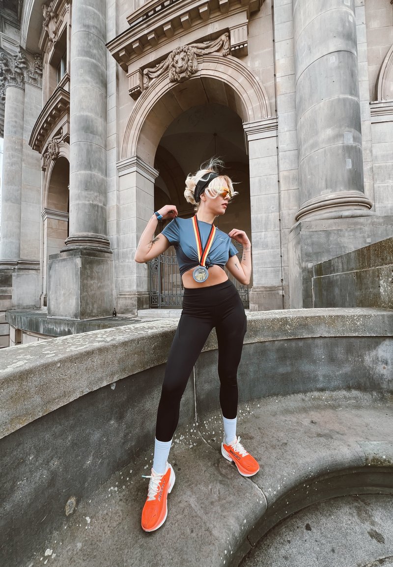 Giovane atleta con una medaglia, scarpe da corsa arancioni, leggings neri e un top corto blu, in posa vicino a storiche arcate e colonne in pietra.