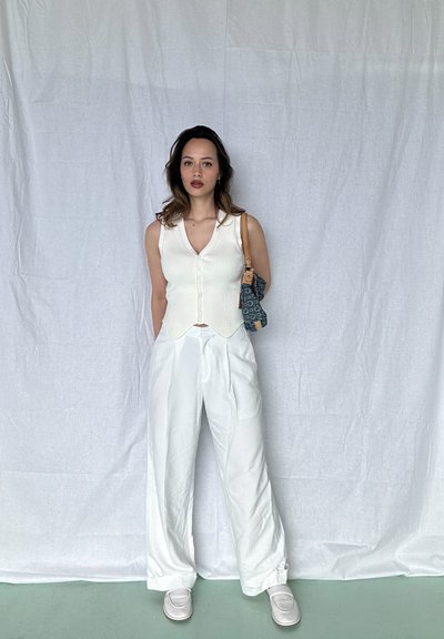 Top blanco de canalé sin mangas con botones, combinado con pantalones blancos holgados. La modelo sostiene un bolso azul con estampado y lleva zapatos de color claro.