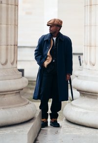 Ναυτικό μπλε παλτό με μπεζ επένδυση, μαύρο πουκάμισο και σκούρο παντελόνι. Καφέ πλεκτό καπέλο και μαύρες μπότες με πορτοκαλί λεπτομέρειες. Πέτρινες κολόνες στο φόντο.