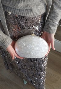 Hvit marmorerte oval clutch med metallisk spenne holdt i hendene, kombinert med en paljettbesatt grått skjørt. Myk tekstur og blank finish.