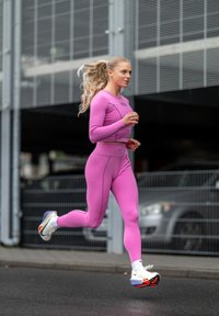 Top sportivo rosa aderente e leggings in tessuto elasticizzato, abbinati a sneakers bianche con accenti colorati, mentre una donna corre sul marciapiede.