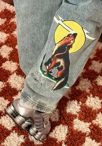 Ben i grå jeans med en farverig ulve-grafik, der hyler mod en gul måne, og sølvfarvede Nike-sneakers på et rødt og hvidt mønstret tæppe.