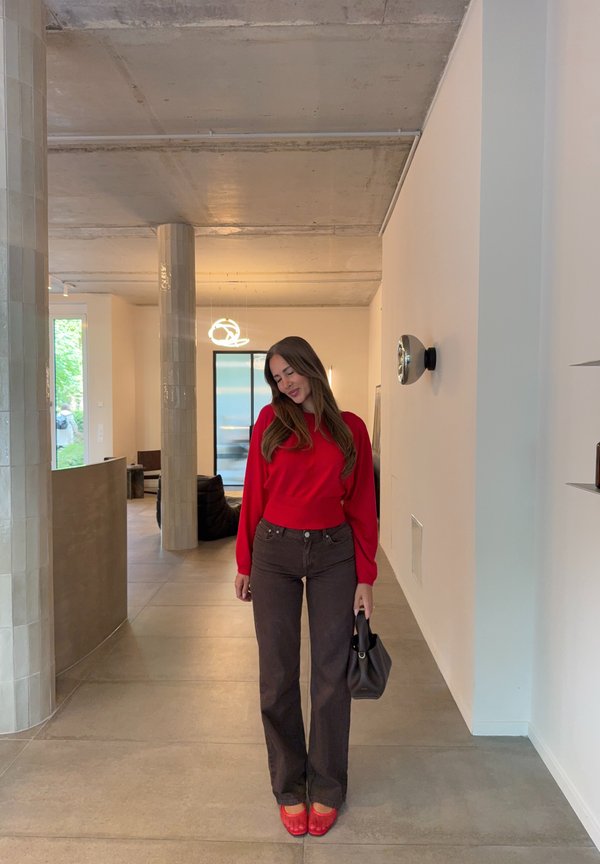 Femme en pull rouge et pantalon foncé debout à l'intérieur tenant un sac noir dans une pièce moderne avec un sol carrelé et un plafond en béton.