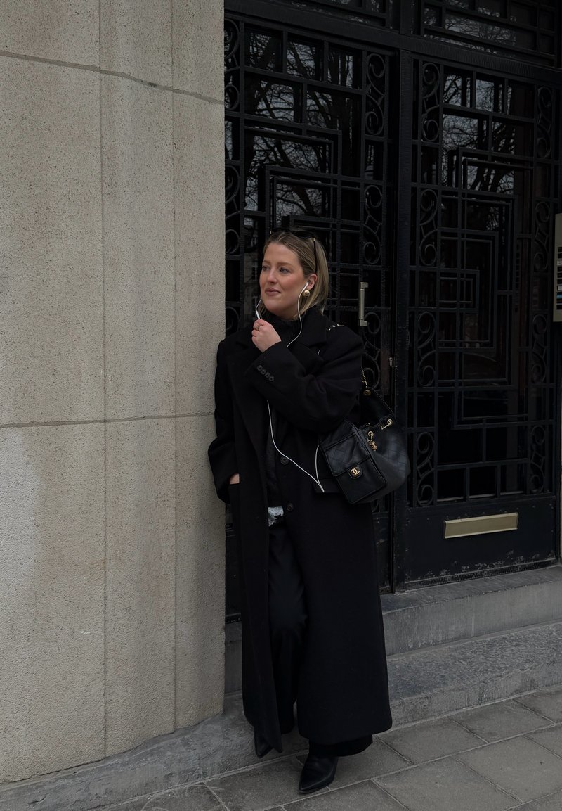 Femme en manteau noir se tient près d'un mur en pierre et d'une porte noire ornée, portant des écouteurs et tenant le câble d'une main.