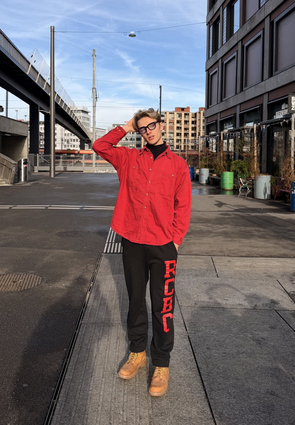 Jeune personne portant un t-shirt rouge et un pantalon noir, debout sur un trottoir urbain, se touchant la tête, avec des bâtiments modernes et des éléments de rue en arrière-plan.