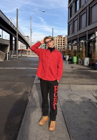 Jeune personne portant un t-shirt rouge et un pantalon noir, debout sur un trottoir urbain, se touchant la tête, avec des bâtiments modernes et des éléments de rue en arrière-plan.