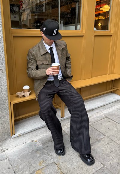 Chaqueta marrón texturizada, camisa blanca, corbata negra, gorra de béisbol negra y pantalones anchos a rayas. Sosteniendo una taza de café, sentado en un banco.