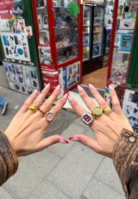 Duas mãos exibem cinco anéis: um de ouro com detalhes em vermelho, um relógio cor-de-rosa, um anel de prata com uma gema vermelha e uma aliança de ouro texturizada.