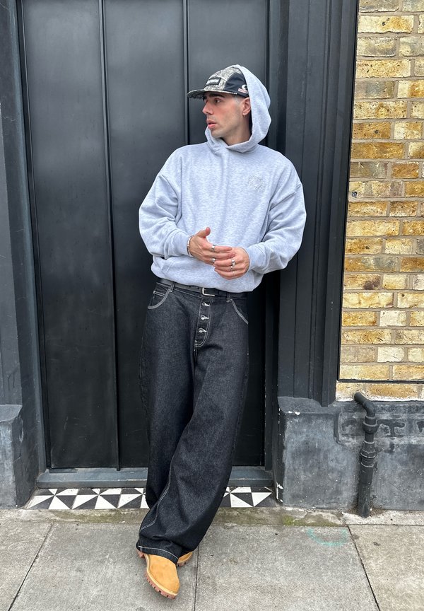 Muž v svetlosivej mikine s kapucňou, čiernej šiltovke, voľných tmavých džínsach a hnedej obuvi sa opiera o čierne dvere rámované tehlovou stenou a pozerá do boku.