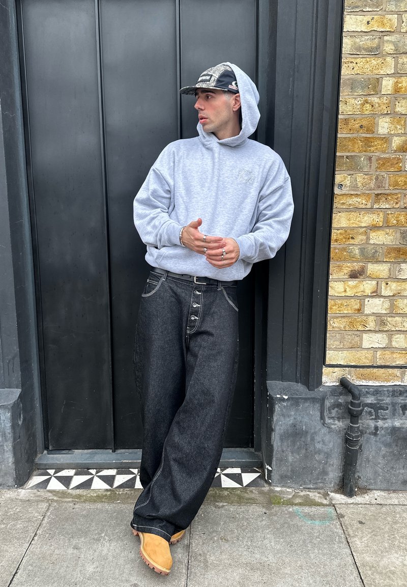 Homme en sweat à capuche gris clair, casquette noire, jean large foncé et bottes marron clair appuyé contre une porte noire encadrée par un mur de briques, regardant sur le côté.