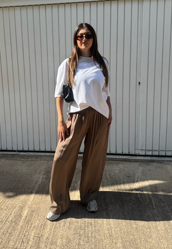 Mujer con camisa blanca de gran tamaño, pantalones marrones de pierna ancha, zapatillas deportivas y gafas de sol, de pie al aire libre frente a una pared blanca.