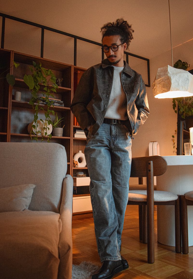 Mann mit Brille und lockigem Haar, der eine Lederjacke, einen Pullover, Jeans und Stiefel trägt, steht in einem gemütlichen Wohnzimmer mit Pflanzen, Bücherregal und sanfter Beleuchtung.