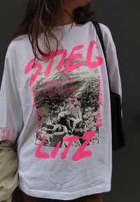 Bílý oversized tričko s černobílým grafickým potiskem a růžovým textem. Materiál vypadá jemně; design obsahuje velký text a květinové akcenty.