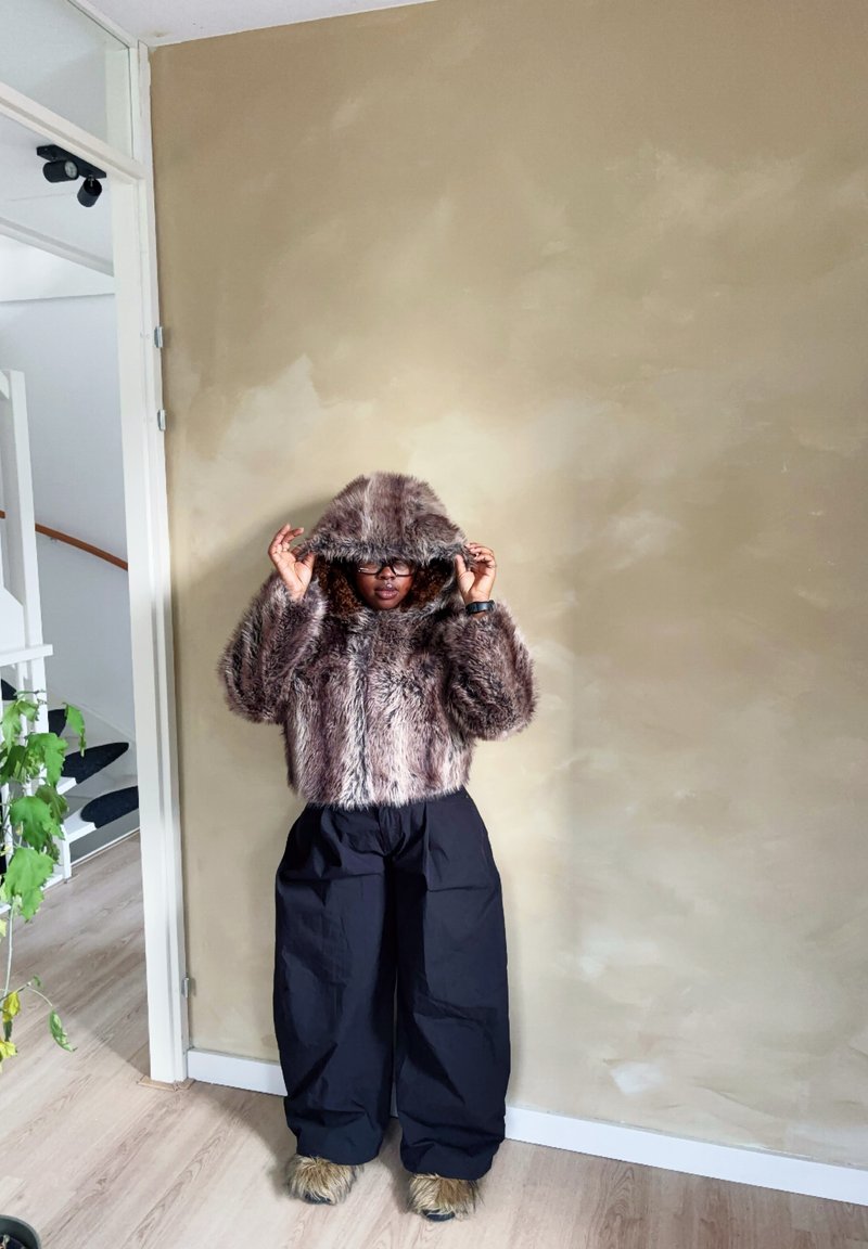 Bruine nepbonten cropped hoodie met een capuchon en zwarte baggy broek. Heeft een textuurstof en een losse pasvorm, tegen een neutrale muur.