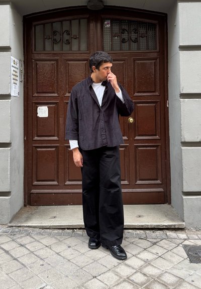 Camisa de botón de pana negra sobre una camiseta de manga larga blanca, combinada con pantalones anchos negros y zapatos negros, frente a una puerta marrón.