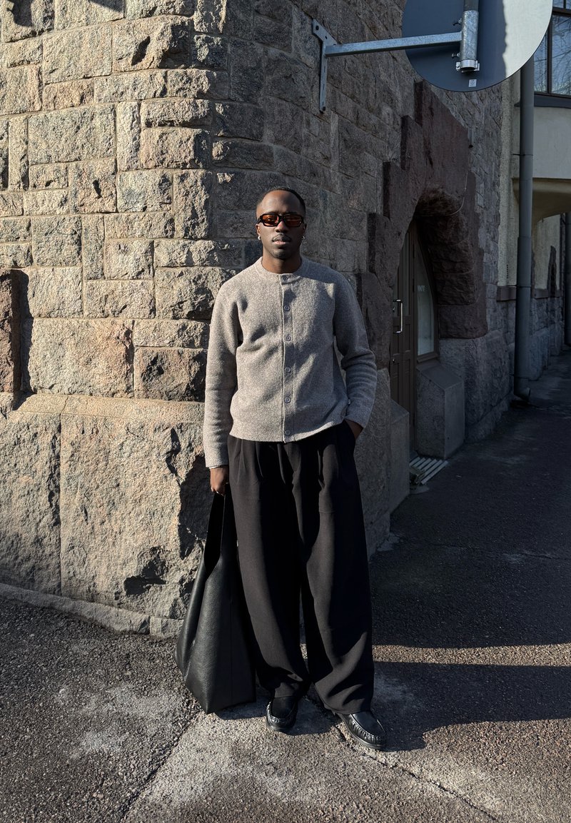 Mann mit brauner Strickjacke und weiten schwarzen Hosen steht an einer Steinmauer und hält in einer urbanen Umgebung eine große schwarze Tasche, im Sonnenlicht mit Schatten.