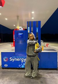 Felpa con cappuccio grigia e pantaloni della tuta; una persona si trova a un distributore di carburante con un sacchetto di snack. Consolle del carburante blu con il logo "Synergy" sullo sfondo.