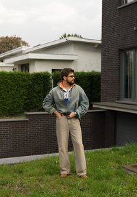 Jaqueta de denim com colarinho laranja, calças largas bege e sapatos castanhos. A cena inclui relva verde e arquitetura moderna ao fundo.