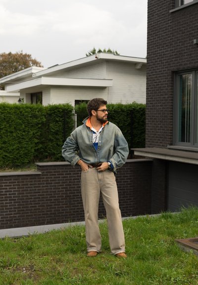 Chaqueta de denim con cuello naranja, pantalones de pierna ancha beige y zapatos marrones. La escena incluye hierba verde y arquitectura moderna de fondo.
