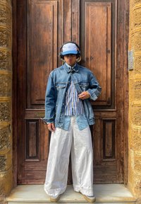 Casaco de ganga por cima de uma camisa azul e branca riscada, combinado com calças largas brancas e sapatos bege. Auscultadores pretos e um boné azul completam o visual.