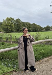 Kvinde i beige trenchcoat og briller drikker iskaffe, står på sti ved en træhegn og en grøn mark under en skyet himmel.