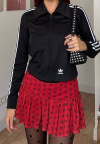 Person som har på seg en svart Adidas zip-jakke med hvite striper, et rødt prikkete skjørt, svarte prikkete strømpebukser, og holder en naglet svart lommebok.