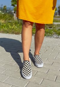 Sapatos slip-on em padrão xadrez preto e branco com solas brancas, usados com uma saia amarela, sobre uma superfície de pavimento texturizada.