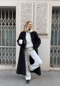 Femme blonde en survêtement gris et manteau noir tenant un gobelet de café à emporter, debout sur une jambe contre un mur texturé avec des fenêtres grillagées.