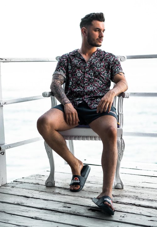 Kurzärmliges, dunkel gemustertes Hemd mit floralem Design, kombiniert mit schwarzen Shorts und schwarzen Badesandalen, sitzt auf einem weißen Holzstuhl.