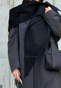 Crni vuneni šal s trokutastim uzorkom prebačen preko sive jakne, upareno s crnom torbom. Ruka drži telefon na kojem su vidljivi bijeli kabeli slušalica.