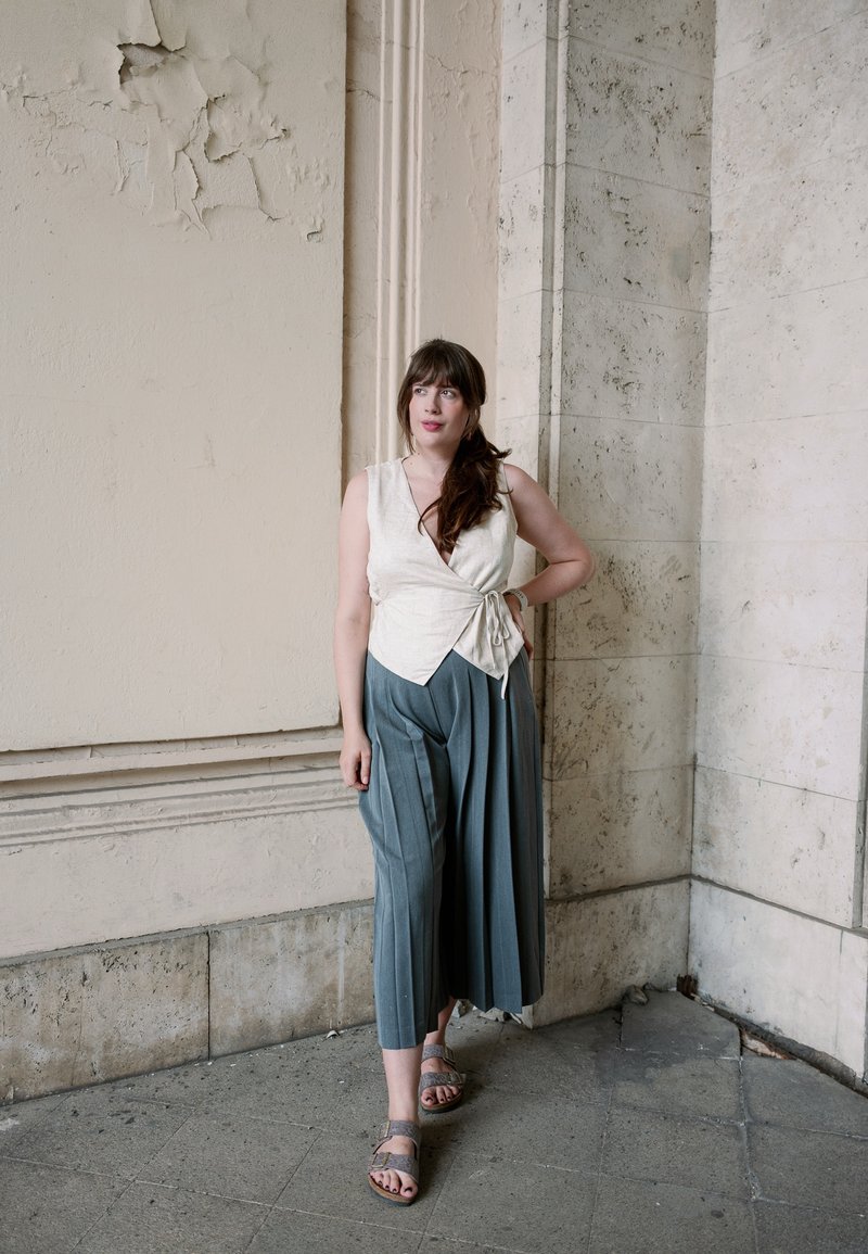 Femme aux longs cheveux bruns portant un haut cache-cœur blanc sans manches et un pantalon gris ample, debout devant des murs en pierre beige texturée.