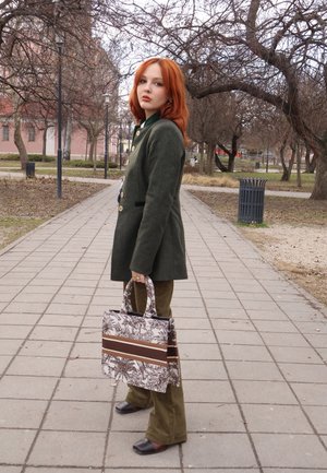 Joven con cabello rojo, abrigo verde y pantalones marrones, de pie en un camino del parque sosteniendo un bolso tote marrón y blanco con estampado.
