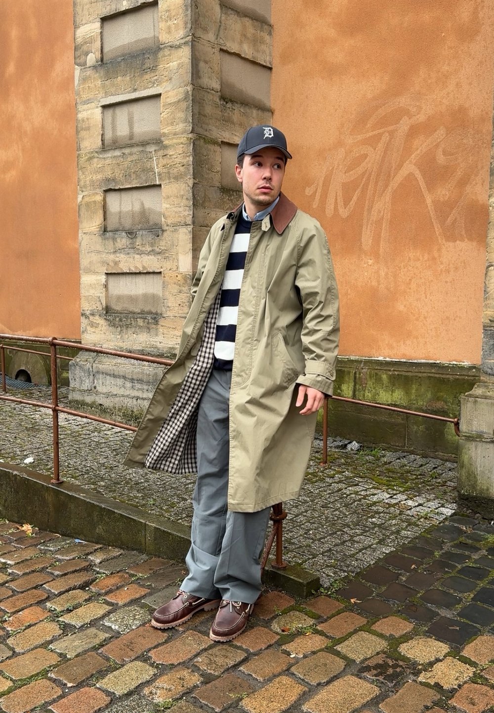 Khaki trench coat with a brown collar, navy and white striped sweater, grey trousers, and brown shoes; standing against a stone wall.