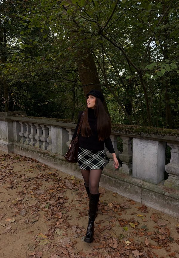 Une femme portant un béret noir, un pull, une jupe à carreaux et des bottes marche le long d’une balustrade en pierre dans un parc verdoyant.