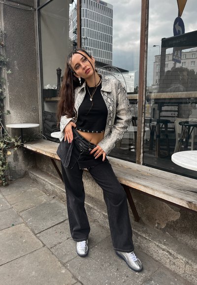 Joven mujer con cabello largo trenzado se apoya en un banco de madera frente a un café, vistiendo una chaqueta de estampado de serpiente, una camiseta de tirantes negra y zapatillas plateadas.