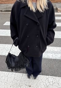 Personne portant un manteau noir croisé, un pantalon foncé et des baskets, tenant un sac à main noir à franges, debout sur un passage pour piétons.
