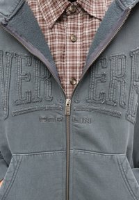 Pilkas užsegamas džemperis su vilnos pamušalu ant apykaklės, siuvinėtu užrašu „EVER BERLIN“. Po juo dėvima ruda languota marškinėliai su sagų užsegimu.