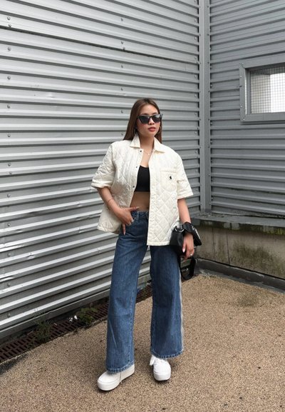 Chaqueta blanca acolchada, top corto negro, pantalones vaqueros de pierna ancha azul, y zapatillas de plataforma blancas, complementados con un bolso negro y gafas de sol.