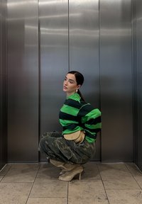 Gestreepte groene en zwarte oversized trui, camouflage cargobroek, beige schoudertas en doorzichtige hoge laarzen in een lift.