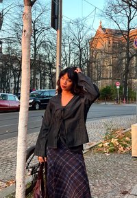 Mørk denimjakke med knyting foran og lange ermer, kombinert med et rutete maxiskjørt i mørke farger. Urbant miljø med trær og parkerte biler.